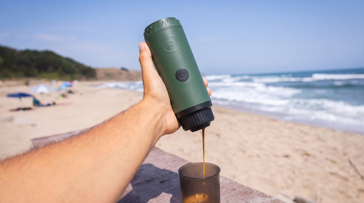 Hand holding a forest green Origin, pouring an espresso into a cup on a table at a sunny beach