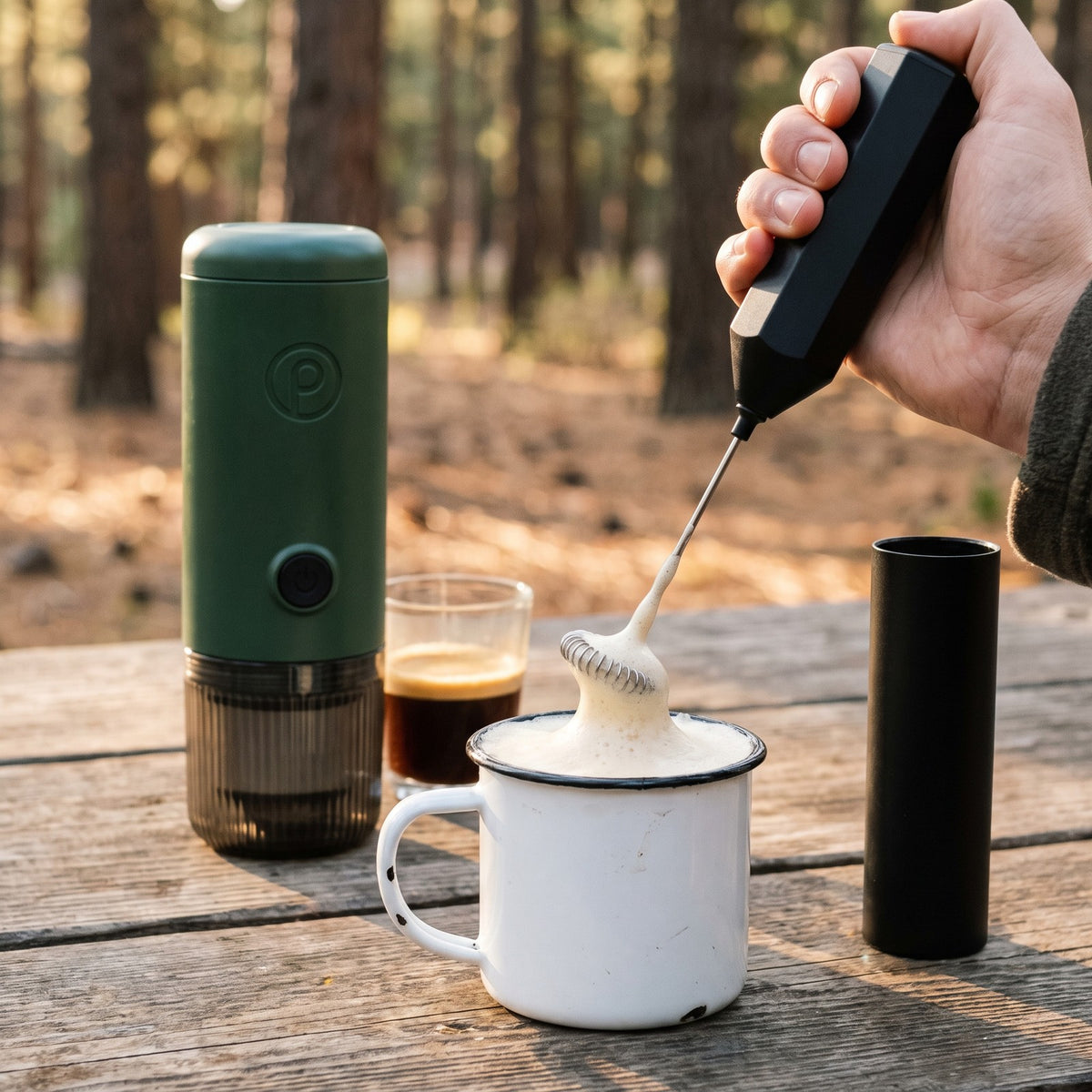 Milk frother whisking milk next to the Forest Green Origin portable espresso maker at camp